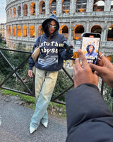 A woman wearing a blue camouflage oversized hoodie with a streetwear style, standing in an urban city setting, holding a bag and wearing sunglasses, exuding a cool and fun vibe.