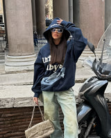 A woman wearing a blue camouflage oversized hoodie stands in a streetwear style pose, with her hands in the pockets. She is also wearing sunglasses and has a stylish, urban background that includes a parked car, highlighting the casual and trendy streetwear vibe.