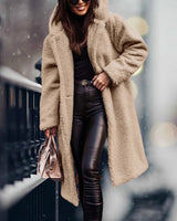 
A woman wearing a long blue teddy coat, characterized by its fluffy texture, suitable for winter fashion. The coat features long sleeves and extends to the knee, offering a cozy and stylish look. The woman is also wearing eyewear, adding a fashionable touch to her outfit.