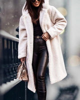 
A woman wearing a long, fluffy blue teddy coat, suitable for winter fashion. She is smiling, with the coat featuring a wide collar, long sleeves, and extending to the knees. The coat is styled to accentuate the waist and has an overall cozy appearance. The woman is wearing eyewear and the coat is available in an XL size, in a creamy white color variant.