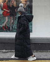 Frau in langem schwarzem Daunenmantel, oversized Maxi Winterjacke, stehend mit grauem Hut und Tasche.