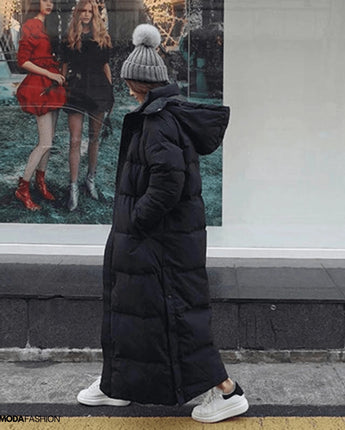 Frau in langem schwarzem Daunenmantel, oversized Maxi Winterjacke, stehend mit grauem Hut und Tasche.