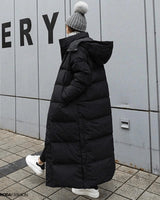 Dame in langem schwarzem Daunenmantel, oversized Maxi Winterjacke, auf der Straße, trägt grauen Hut und Stiefel, Fellbesatz an der Kapuze.