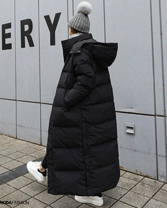 Dame in langem schwarzem Daunenmantel, oversized Maxi Winterjacke, auf der Straße, trägt grauen Hut und Stiefel, Fellbesatz an der Kapuze.